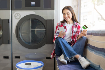 A young woman sits comfortably on a bench in a laundromat, using her smartphone while holding a payment card. She smiles as she waits for her laundry to finish.