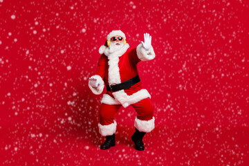 Full body size photo of pensioner old man grey beard chilling middle dance floor wear santa costume gloves coat belt sunglass headwear earphones black boots isolated red color background