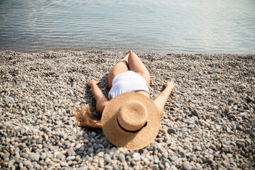 A woman is laying on the beach wearing a straw hat. She is wearing a white bikini top and white bikini bottoms.
