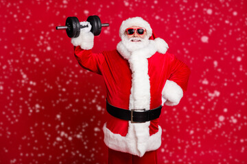 Photo of pensioner old mad man white beard lift heavy dumbbell easily shiny smiling confident self-assured hand hip wear santa x-mas costume sunglass cap isolated red color background