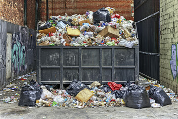 A cluttered dumpster overflowing with various types of waste, surrounded by litter in an urban alleyway.