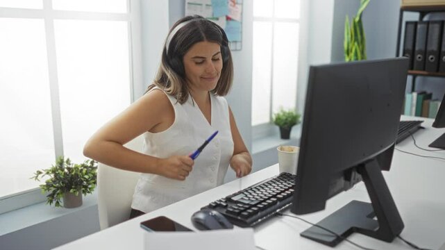 Young woman wearing headphones working in an office, engaging with a keyboard and enjoying her music while holding a pen.