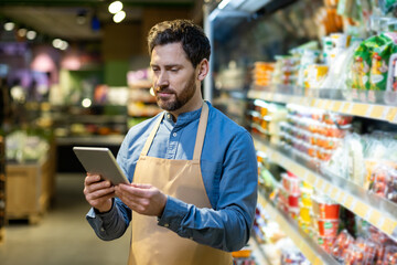 Supermarket employee wearing apron uses tablet in grocery aisle. Man manages inventory with modern technology, demonstrating efficiency and focus in retail environment.