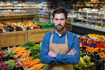 Portrait of confident male employee in grocery store displaying fresh produce. Worker wearing apron in market environment with carrots, radishes, and peppers showcasing healthy lifestyle food concept