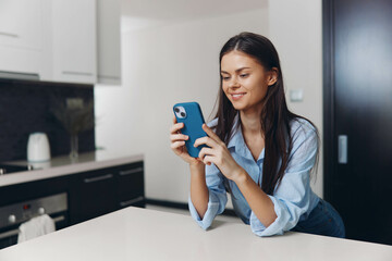 Happy woman sitting at kitchen table looking at phone and smiling at camera, concept of modern technology and communication