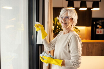 Smiling senior woman cleaning window with detergent at home.