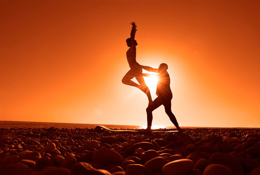 Silhouette of fit sporty couple practicing acrobatic yoga with partner together on the beach. Female acrobat balancing on feet of male partner in fylfot shape. pair doing acro yoga on sea at sunrise.