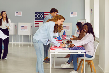 Side view portrait of smiling happy young girl american citizen registering at polling station with American flags on election day. USA woman voter standing at vote center and getting ballot paper.