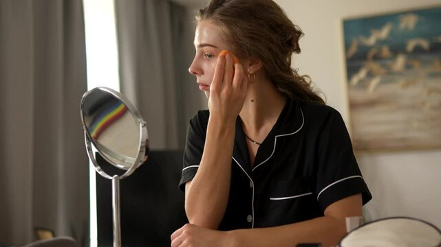 Young woman doing makeup, looking at mirror sitting in cozy room. Beautiful woman making herself beautiful indoors. Beauty, makeup concept.