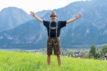 An Austrian farmer in national dress stands against a backdrop of mountains and a small town, arms spread wide.