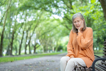 Senior woman suffering from toothache, sitting outdoors on a bench with pained expression. She is holding her face, showing discomfort. Natural green environment adds tranquility contrast to her pain