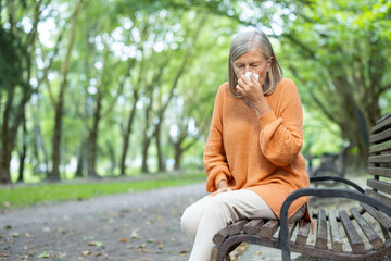 Senior woman in orange sweater sitting on park bench sneezing into tissue. Outdoor setting during...