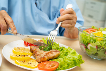 Asian senior woman patient eating pork chop stake and vegetable salad for healthy food in hospital.