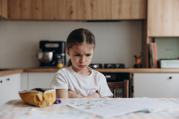 upset schoolgirl sitting at home at the table doing homework from school
