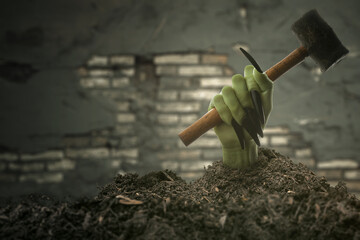 Witch hand with long black nail coming out from grave ground holding a hammer with brick wall background. The hand appearing to be a evil, satan, zombie or a monster hand