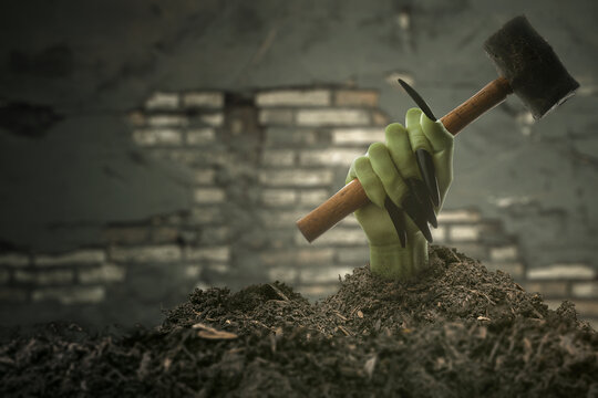 Witch hand with long black nail coming out from grave ground holding a hammer with brick wall background. The hand appearing to be a evil, satan, zombie or a monster hand
