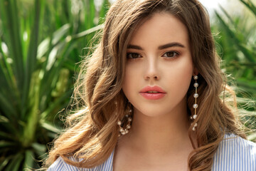 Naklejka premium Young beautiful woman with perfect skin and big pearl earrings and curly hair front of greens plants. Close up, selective focus.