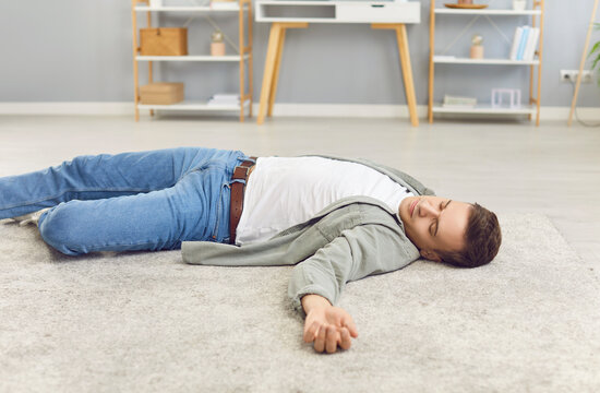 Portrait of a young unconscious man in casual clothes lying alone on floor in living room at home with heart attack or cardiac arrest. Male person fainted indoors. Health and first aid concept.
