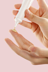 Womans hands gently applies a nourishing skin serum from a sleek white tube onto their hand, set against a soothing soft pink background. This serene scene beautifully emphasizes the importance of ski