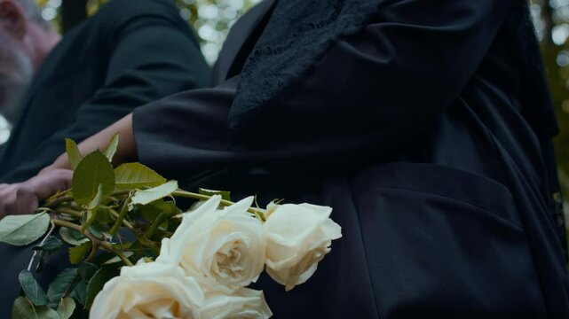 Close up tilt down shot of woman dressed in black sitting with bouquet of roses on her lap and holding hand of husband during visit to cemetery