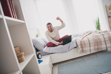 Full length photo of handsome young guy sit bed netbook winning dressed sleepwear wake up spacious house indoors bedroom