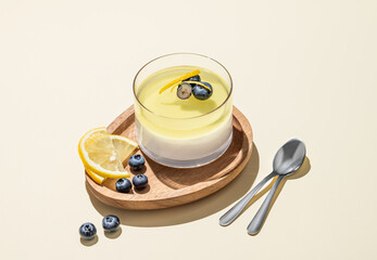 Lemon panna cotta with blueberries in a glass on a wooden board on a yellow background with fresh berries, spoons and shadow.