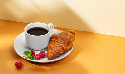 Cup of coffee and fresh croissant with raspberries on a white plate on an yellow background with shadow.