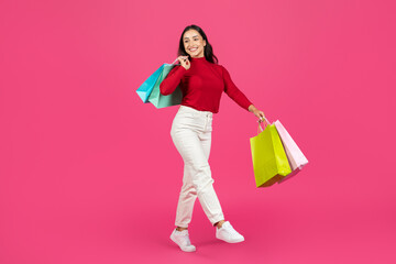 A cheerful woman dressed in a red sweater and white pants holds multiple shopping bags in bright colors. She poses playfully, showcasing her excitement for shopping.