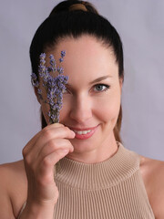A smiling white woman with her hair in a ponytail, holding a small bouquet of lavender close to her face, partially covering one eye. She is wearing a beige sleeveless top