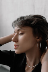 Obraz premium Woman elegantly poses indoors, softly illuminated by natural light. She gently holds her hair back, displaying a serene expression and minimalist jewelry that enhances her beauty
