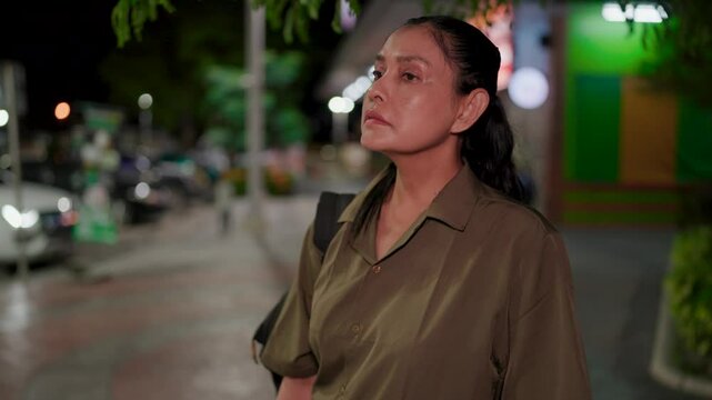 A front view of a middle-aged woman using her phone at night, waiting for something or someone as she looks around, checking her phone and making a call, looking around as she wait. Depicting the soli