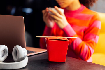 Close-up of red box with Asian food and chopsticks on table. Lunch snack at freelancer job at home. Young African woman is drinking tea and working on laptop.