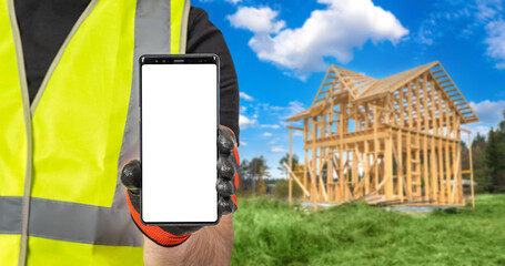 Mobile phone in hand of builder. Smartphone with blank white screen. Cropped man demonstrates phone. Place for advertising construction company. Worker near wooden frame of house under construction.