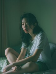 Obraz premium Asian woman in white t-shirt, sitting on bed with hands clasped, looking to side. Blurred background creates intimate atmosphere.