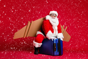 Full length photo of retired childish grandpa white beard ride suitcase reactive jetpack pilot ascending sky wear santa x-mas costume spectacles headwear isolated red color background