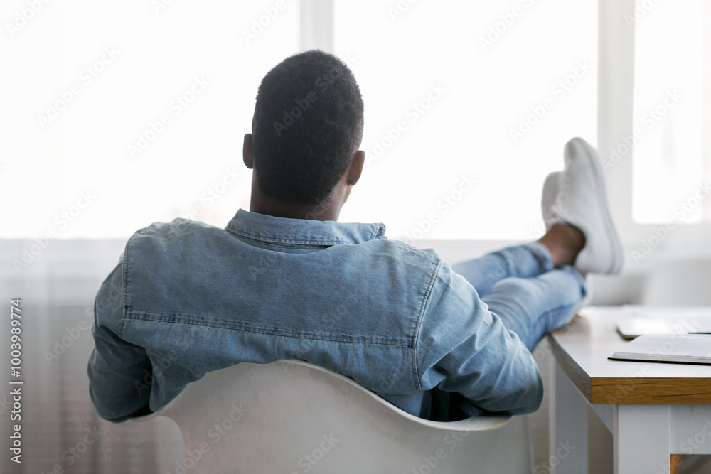 Poster idle time concept. rear view of lazy african american employee sitting with his legs on work desk an