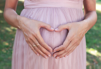 Young pregnant woman in pink clothes on green background. Woman in 9th month of pregnancy. Woman touches her belly with her hands and shows heart sign. Concept of expecting a baby.