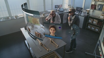 Fototapeta premium Advanced archaeological lab: Two archaeologists in VR headsets study skeleton remains using augmented reality. 3D animation of holographic display with AI voice assistant and information web browser.