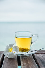 Cup of green tea with ginger servered with plumeria frangipani flower front of at sunset light on wooden tabletop. Sea view.