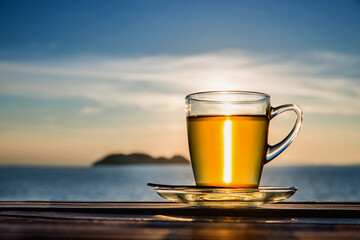 Cup of amber green tea  on wooden tabletop shining in the sunset light. Sea view.