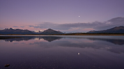 Fototapeta premium A tranquil scene unfolds at Lago Roca, Patagonia, as the moon illuminates the serene lake and reflects the majestic snow-capped mountains against a breathtaking twilight sky.