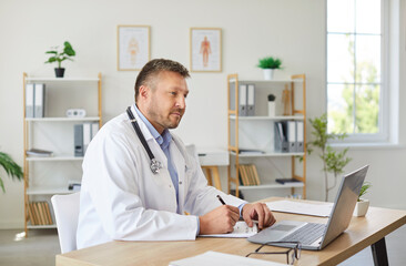Portrait of confident mature male doctor sitting at the desk and working in office looking at...
