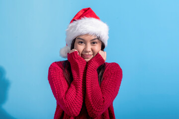 Asian woman dressed in a red knit sweater and wearing a Santa hat smiles sweetly while resting her fists against her cheeks. The background is a solid light blue, creating a cheerful holiday scene