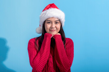 Asian woman dressed in a red knit sweater and wearing a Santa hat smiles sweetly while resting her fists against her cheeks. The background is a solid light blue, creating a cheerful holiday scene