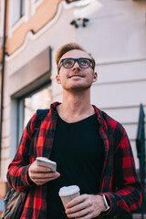 Smiling guy with smartphone and hot beverage looking up