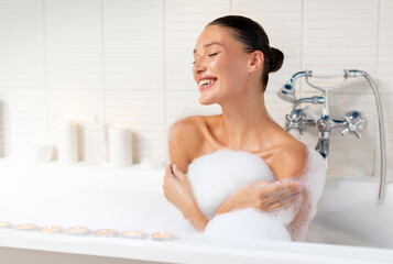 Fototapeta premium Happy woman taking relaxing bath with foam at home, enjoying warm water in bathroom interior in morning, copy space