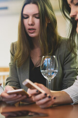 Charming female bloggers checking notification on smartphone gadgets during friendly meeting in cafe interior for drinking white wine, caucasian women reading news in social networks via cellphones