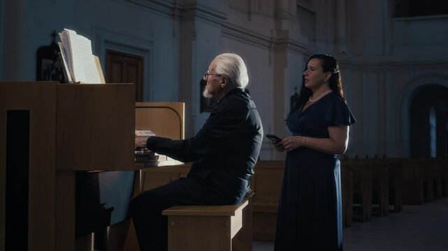 Female opera singer performing to accompaniment of organ played by elderly musician in empty cathedral