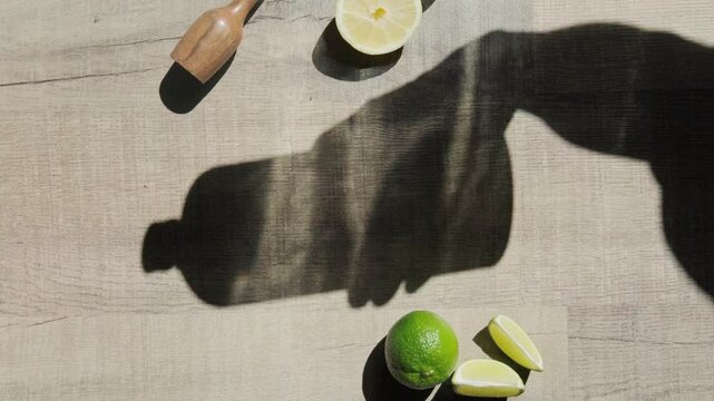 An arrangement of lemons and limes with a grater and muddler on a wooden surface. Sunlight casts a shadow of a bottle, creating an artistic composition ideal for a culinary or beverage preparation the