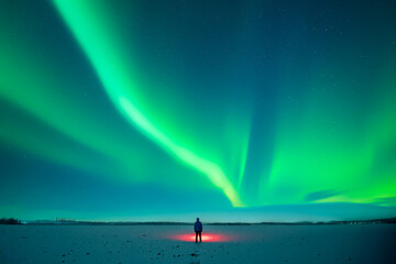 Tourist with red flashlight standing on snowy field against the backdrop of incredible starry sky with Aurora borealis. Amazing night landscape with Polar lights. Northern lights in winter field